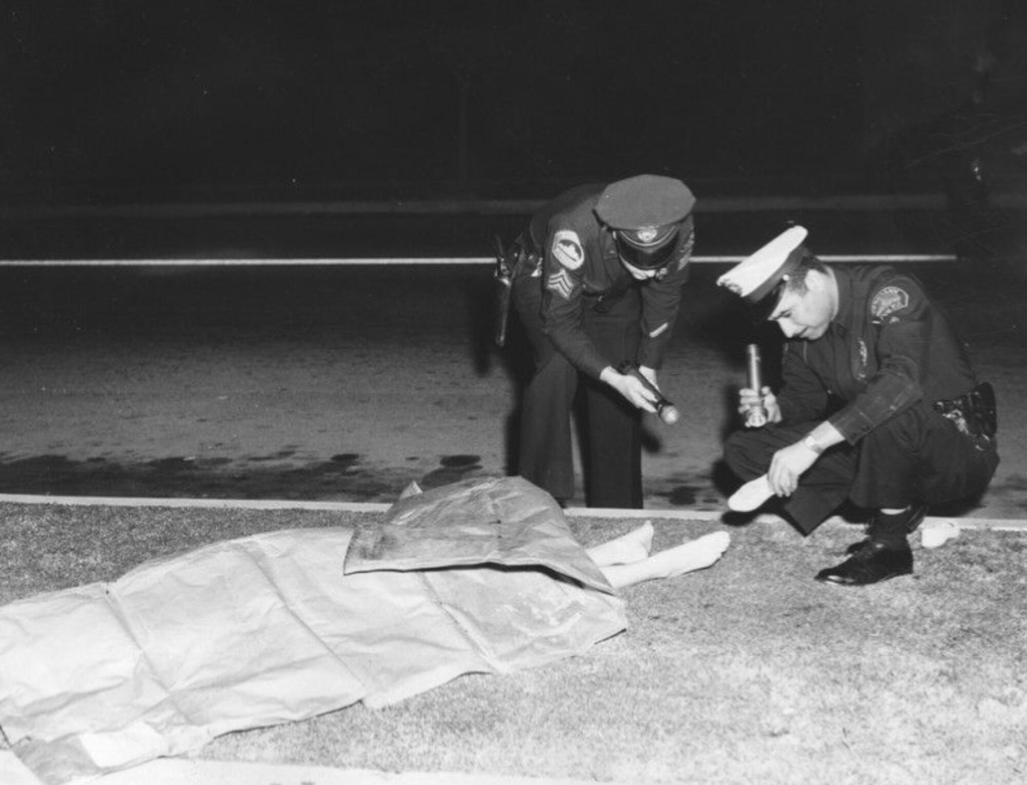Two police officers inspecting a body on a grassy area outdoors. Most of her body is covered by cloth but her feet visible.