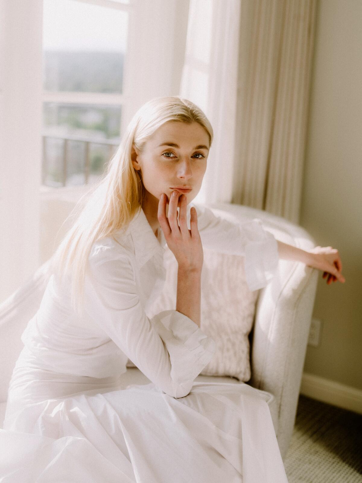 Elizabeth Debicki wears a white dress and sits in a white chair for a portrait.