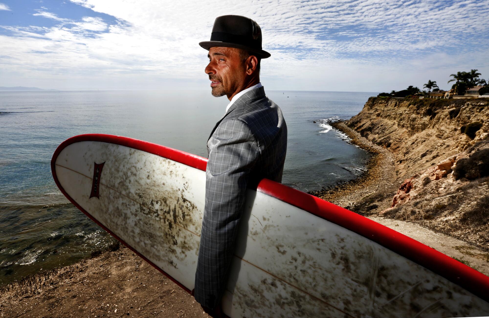 A man in a hat and suit jacket holds a surfboard under his left arm.