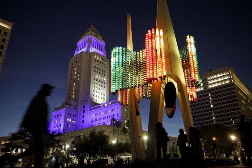 People walk past the Triforium illuminated with LED lights as part of a restoration effort as City Hall stands illuminated in Dodgers blue at night on Friday, October 26, 2018 in Los Angeles, Calif. The six-story, 60-ton public sculpture at Fletcher Bowron Square in the Los Angeles Mall Civic Center complex was created in 1975 by artist Joseph Young, however technical problems plagued the music and sound responsive sculpture. (Patrick T. Fallon/ For The Los Angeles Times)