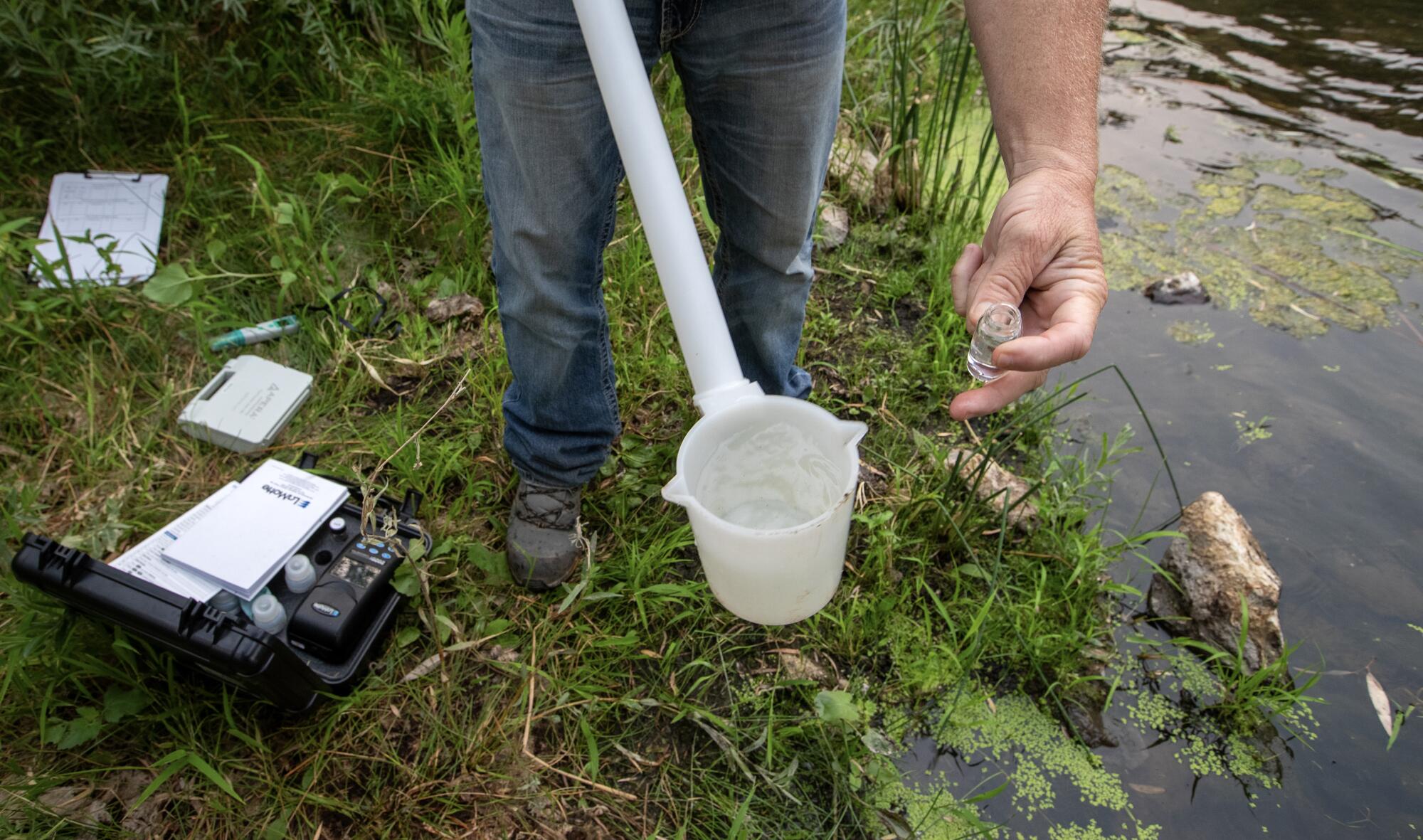 A person on the bank of a body of water holds a long handled bucket in one hand and a small vial in the other.