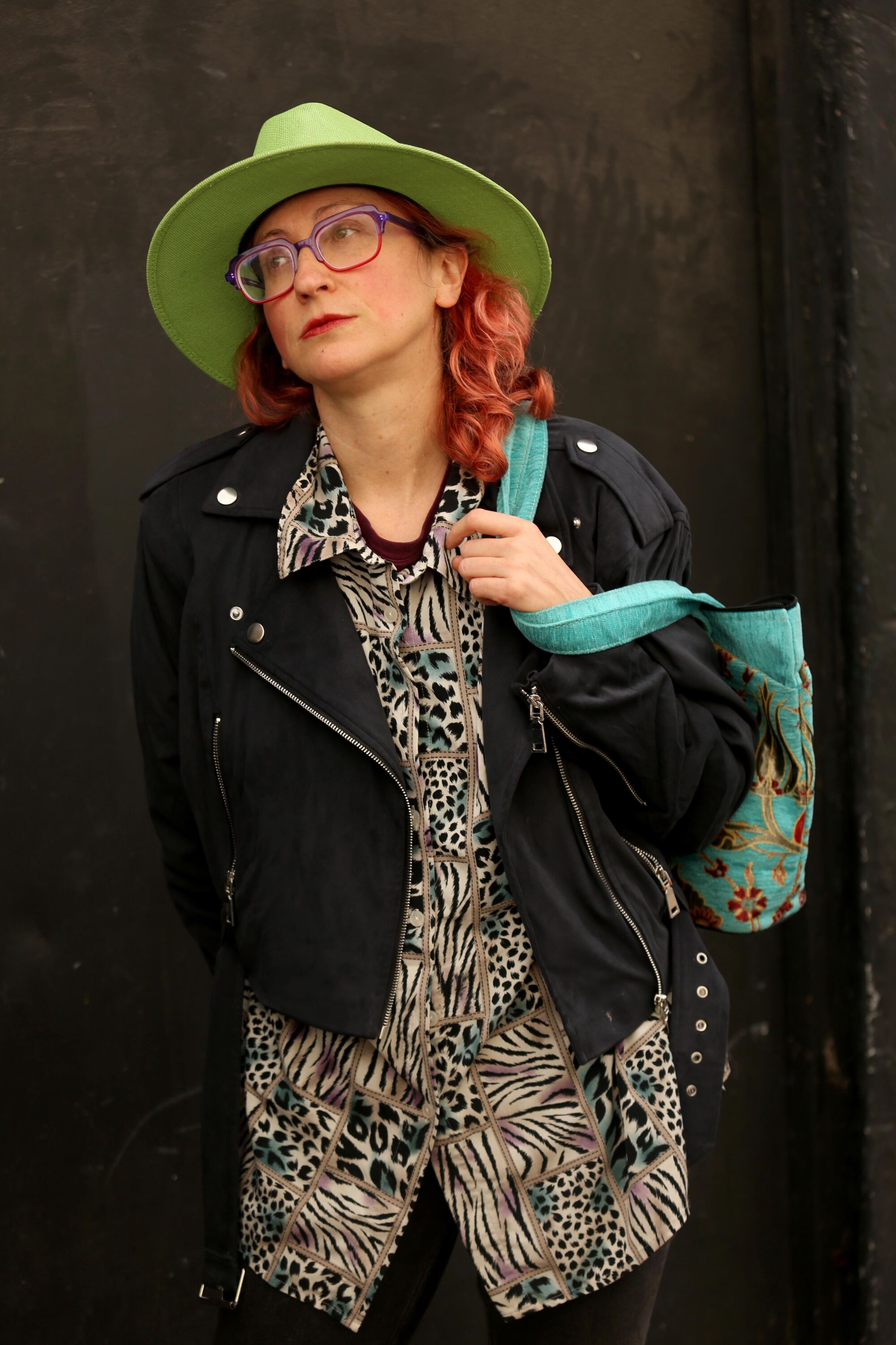 Rachel Mason in a black jacket, patterned shirt and green hat looks to the side while holding a colorful tote bag.
