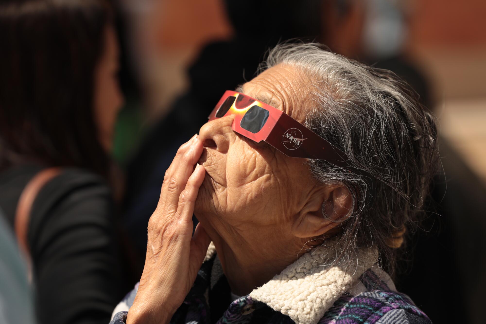A woman in eclipse glasses looks up and puts her hand to her face.