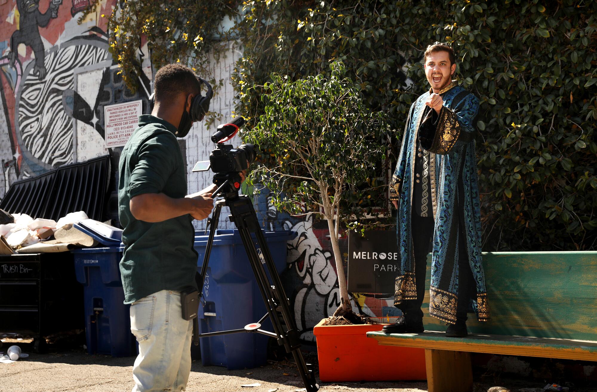 An actor in a costume robe performs a monologue on top of a bench while another man films.