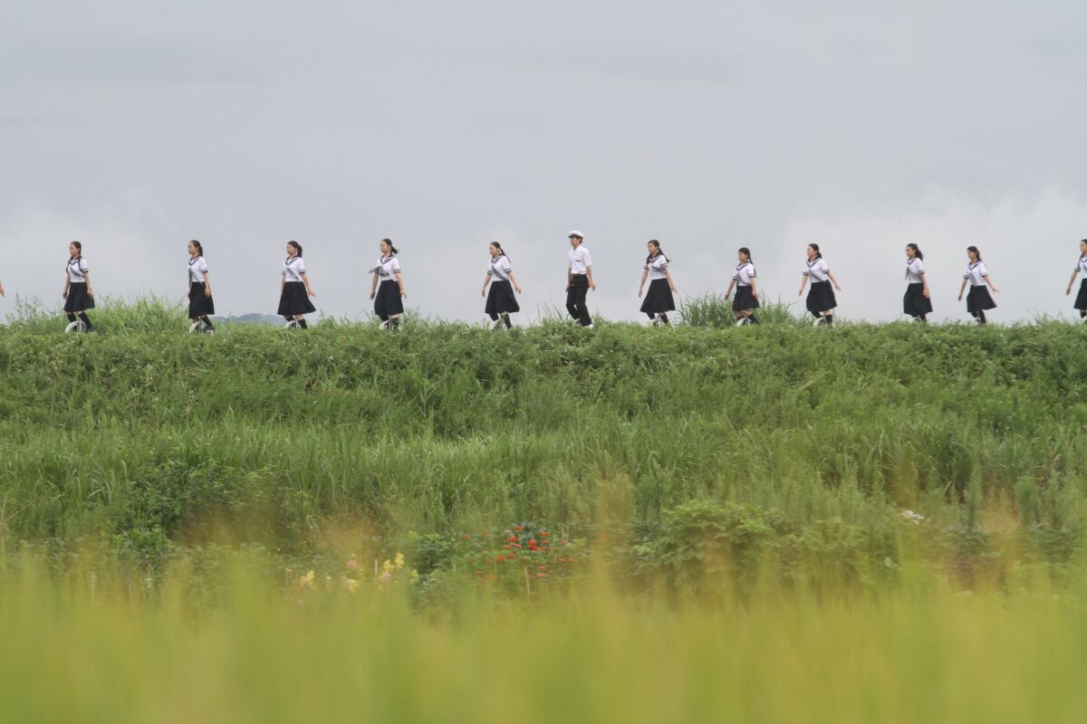 Young women and a man march single file across a grassy berm  