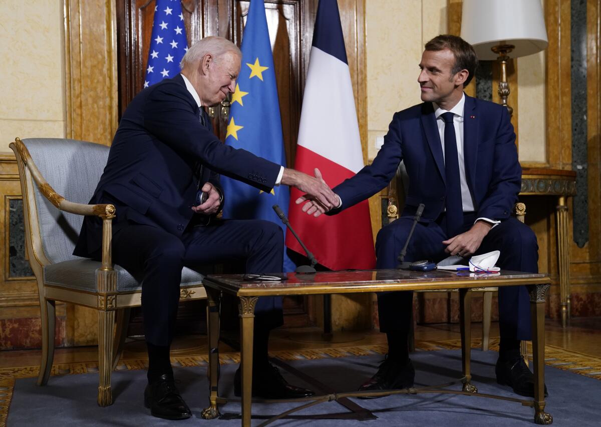Two men in shake hands while sitting in chairs at a coffee table. Three flags are behind them