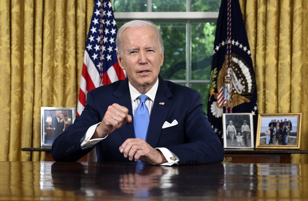President Biden sits at his desk and speaks with golden curtains and flags behind him.