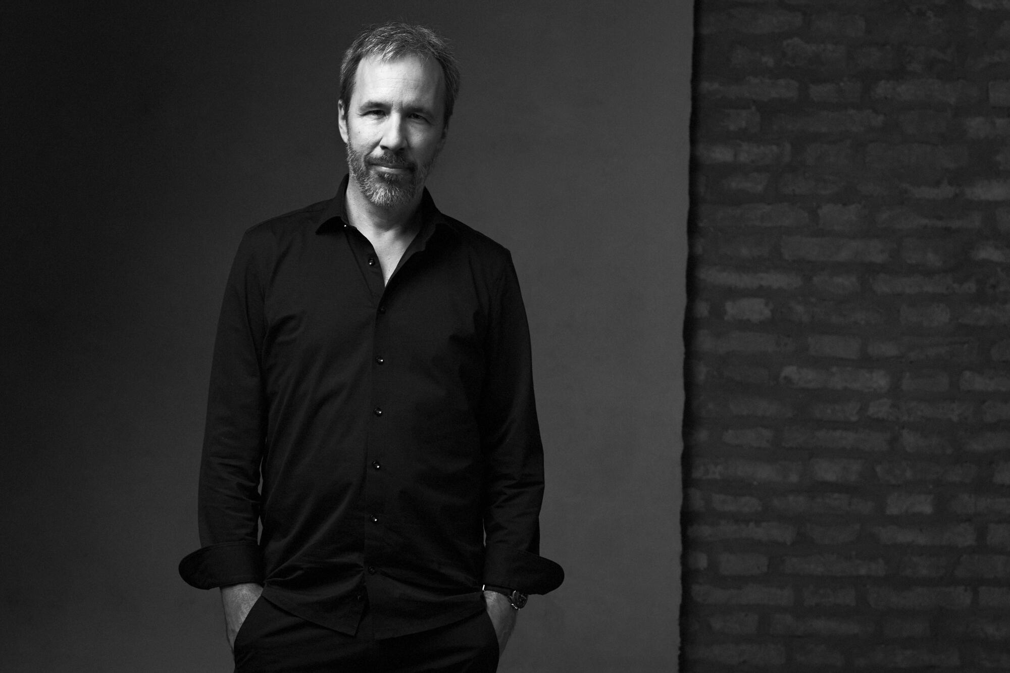 A portrait of filmmaker Denis Villeneuve, all in black, standing with his hands in his pockets.