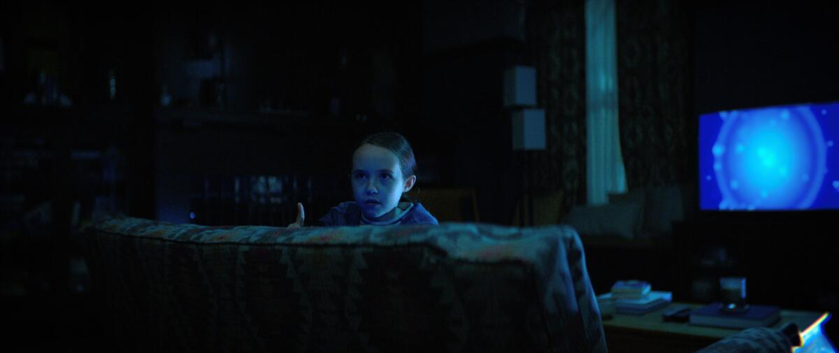 A young girl looks over the couch she's sitting on, with a blue TV screen behind her illuminating the room.