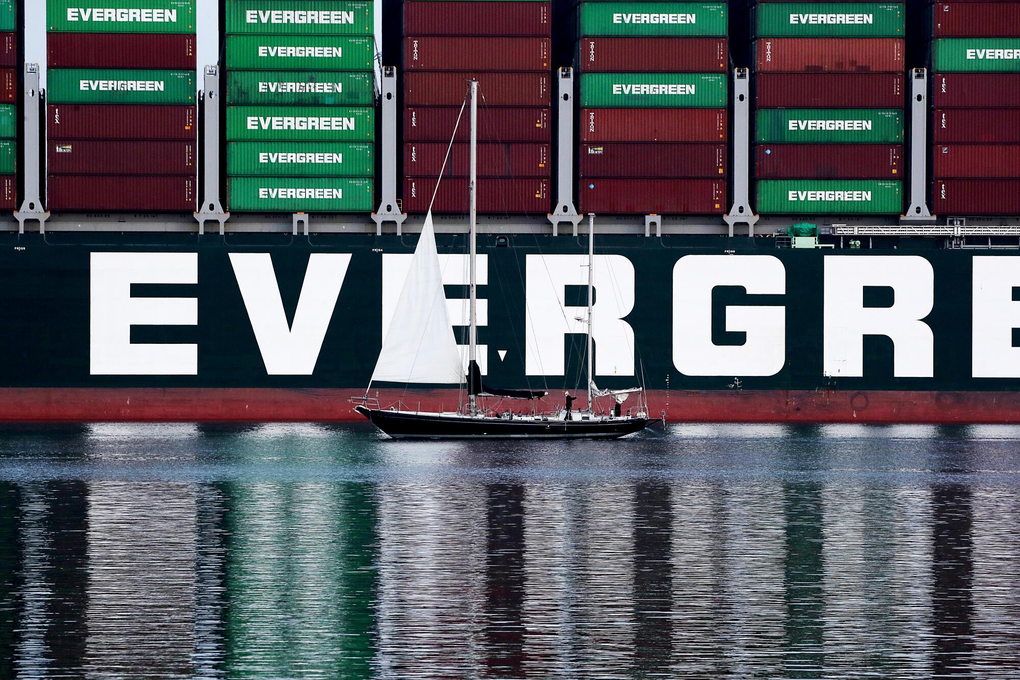 A pleasure boat is dwarfed by a ship and stacks of cargo containers 