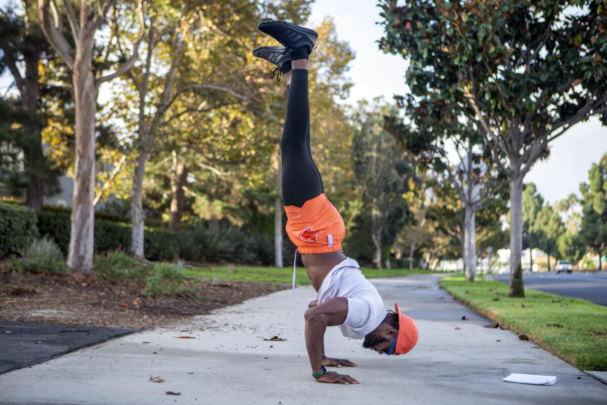 A young Black man exercises, doing a hand stand on a sidewalk lined with green trees.