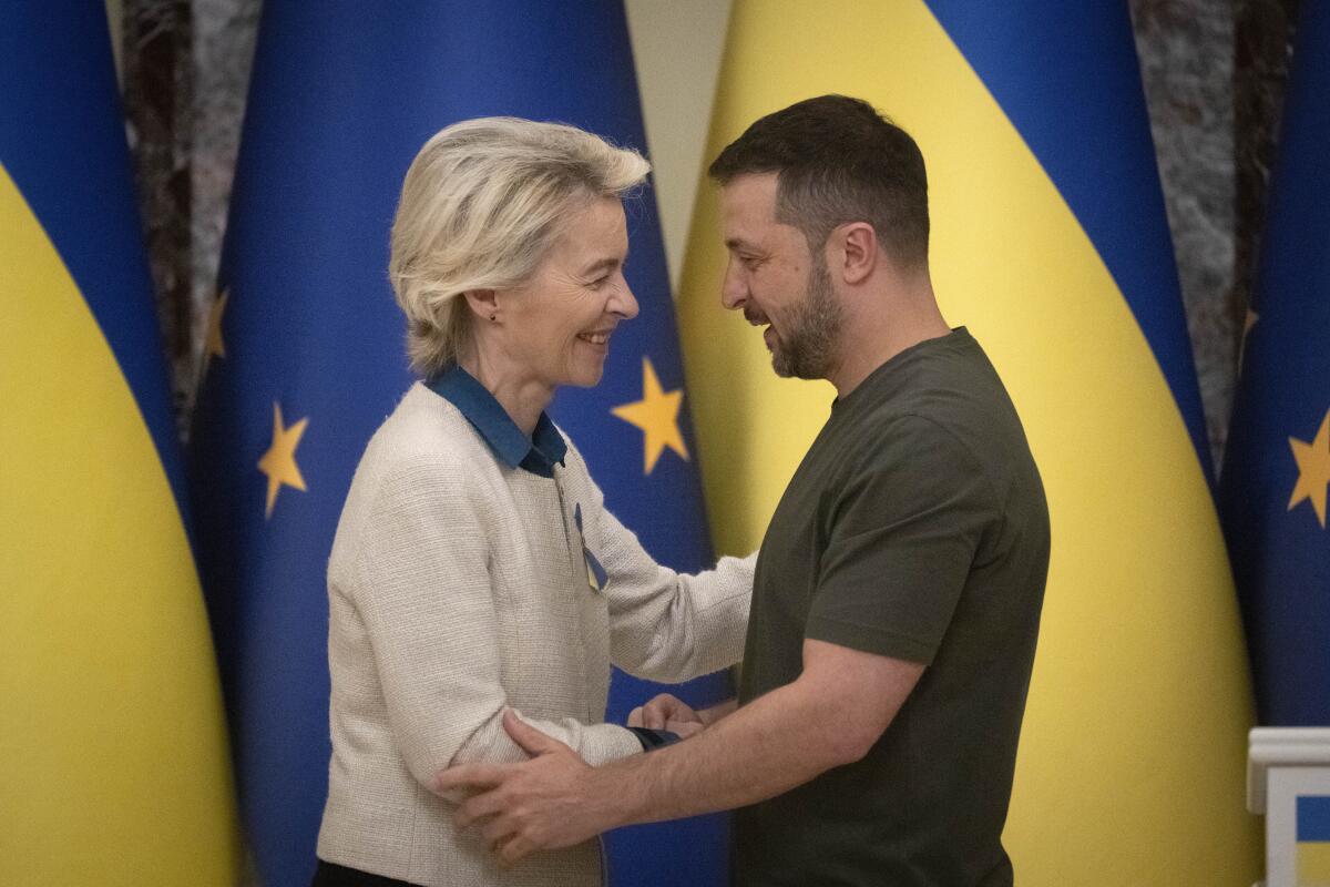 A man in an olive T-shirt and a woman in a shirt and sweater greet each other with outstretched arms in front of flags