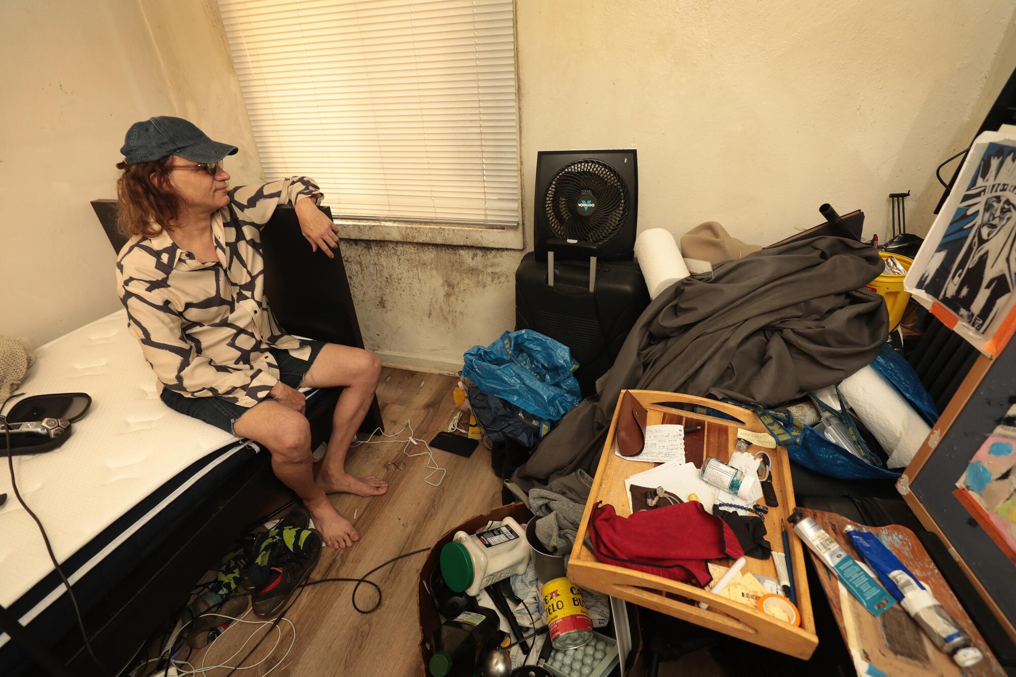 A person sits on a bed in a cluttered room and looks at mold under the window