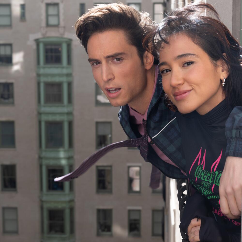 A man and a woman lean against the railing of a window of a tall building.