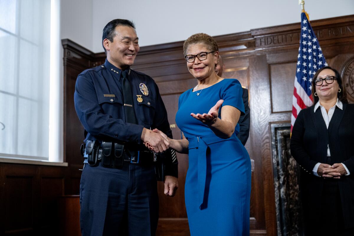 A man in dark police uniform, left, and a woman wearing glasses and a blue dress smile while shaking hands