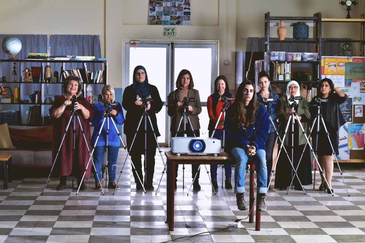a bunch women stand behind cameras behind a projector in a room with linoleum floors 