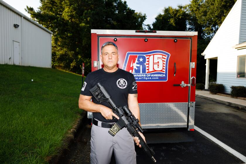 A man stands in front of a red truck, holding an AR-15 assault rifle.