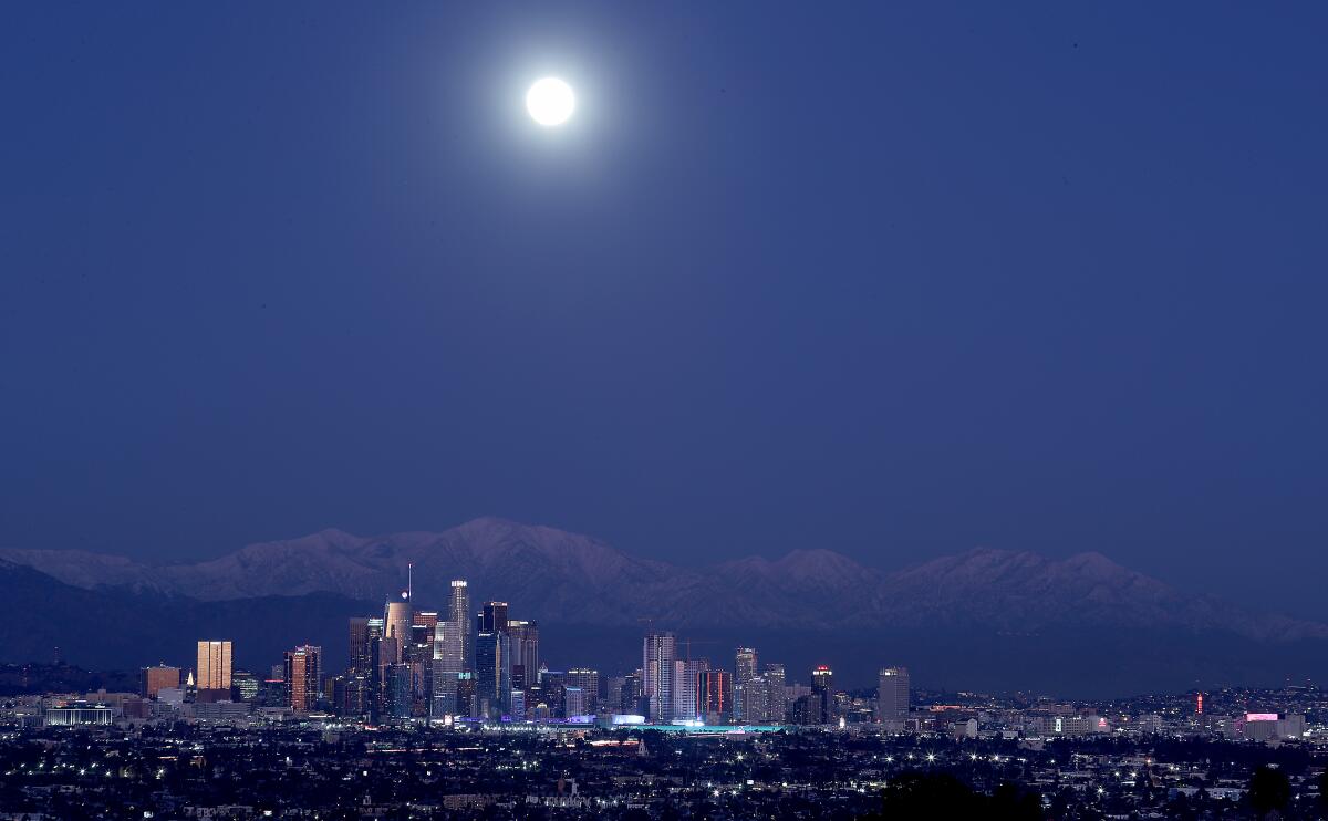 A full moon rises, with the downtown L.A. skyline and the snow-capped San Gabriel mountains visible