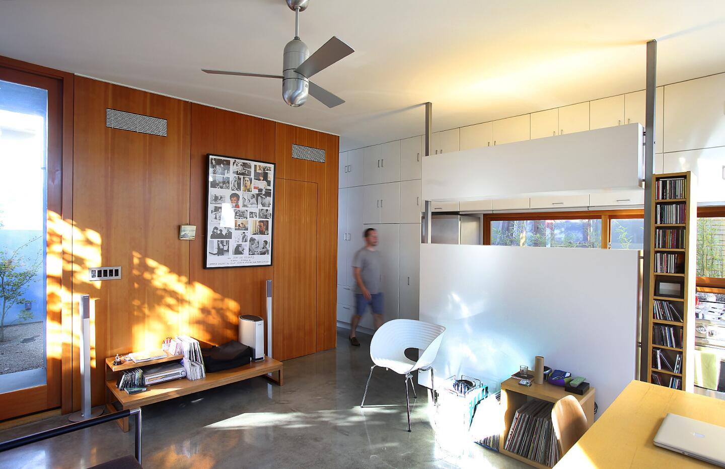 Tenant Joshua Selsky walks toward the kitchen, hidden behind a white room divider that doubles as a projection screen. Natural light spills into the main living-dining area from three sides. Douglas fir wall panels on the left keep media equipment tucked out of sight. - by Emily Young