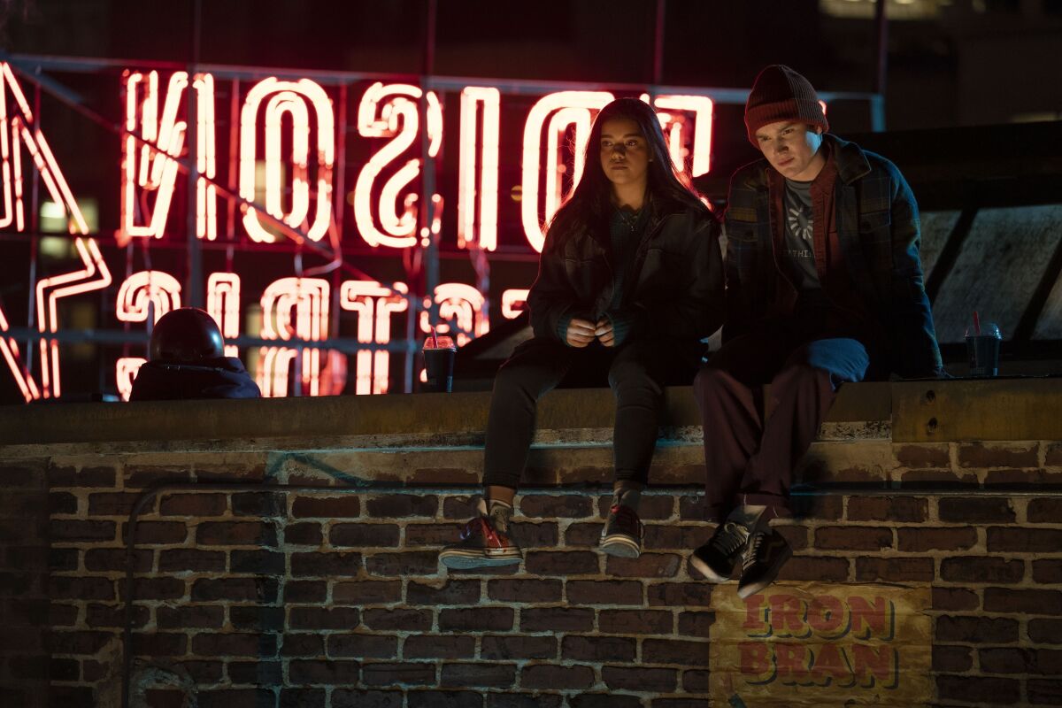 Two teens sitting on a rooftop with a neon sign behind them.