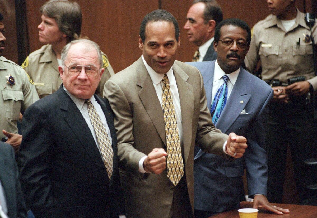 O.J. Simpson reacts with clenched fists while flanked by two men in suits and law enforcement officers stand behind him.