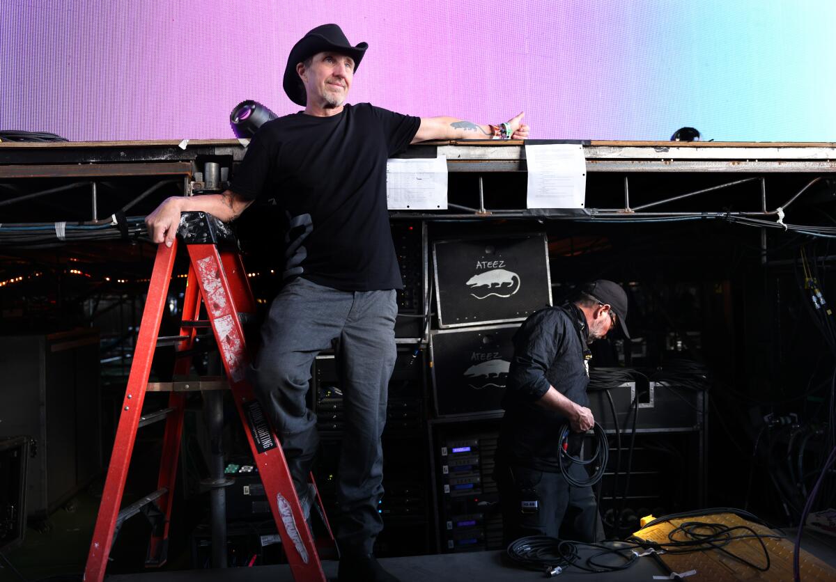 Man in black cowboy hat standing on a ladder near sound equipment.