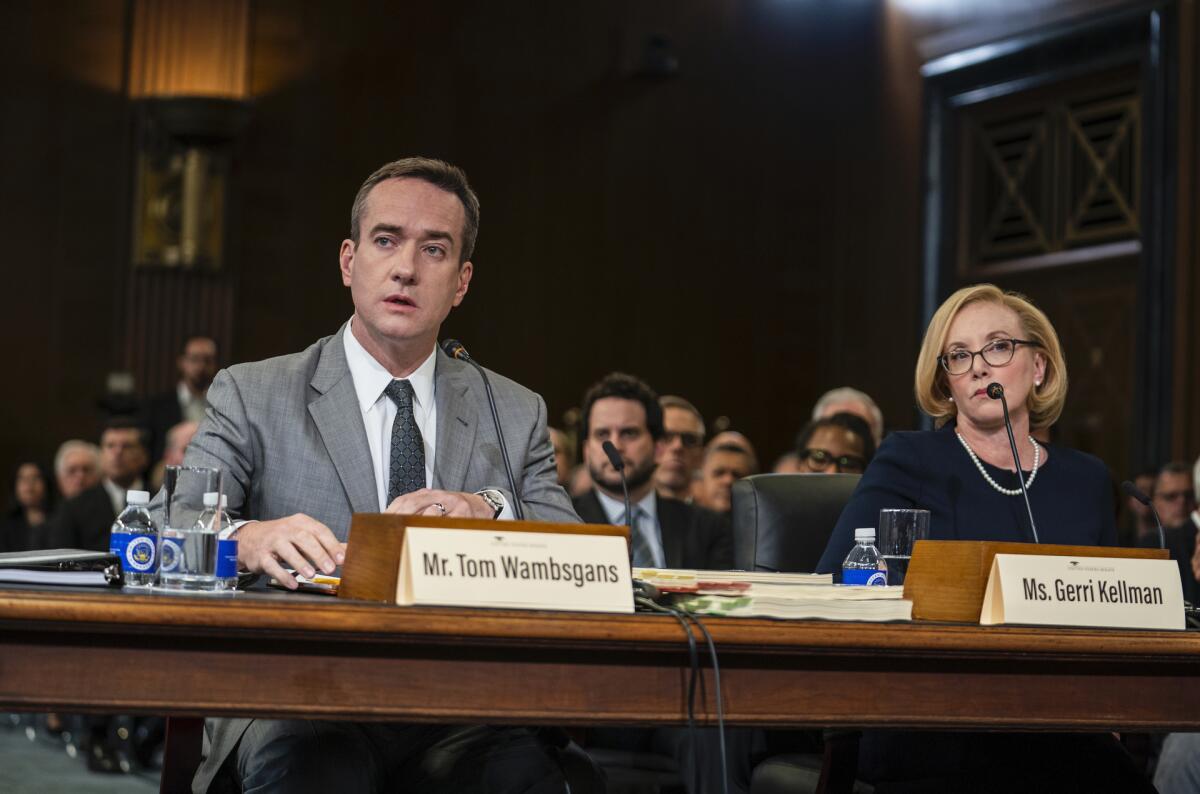 A man in a gray suit and a woman in a black dress and glasses sit at a wooden table with name cards during a Senate hearing.