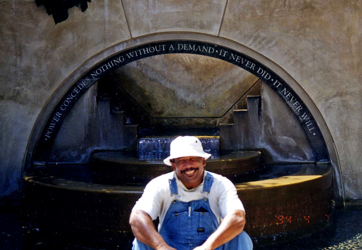 Eric Priestley sits in front of a fountain that reads, "Power concedes nothing without a demand"