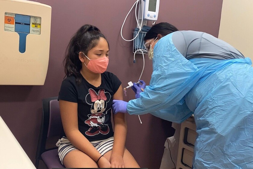 A masked girl wearing a black Minnie Mouse T-shirt, who is seated, gets a shot in the arm from a health worker 