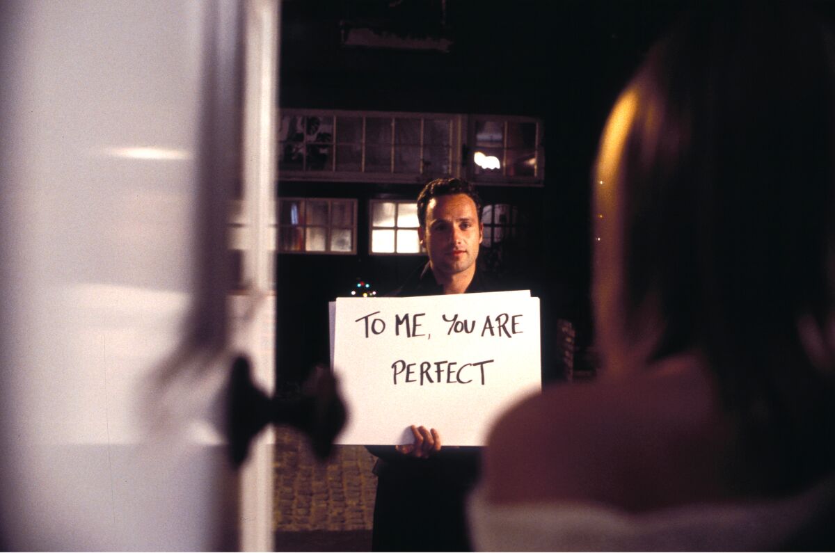 A man holds a cue card reading "TO ME YOU ARE PERFECT" at a woman's front door.