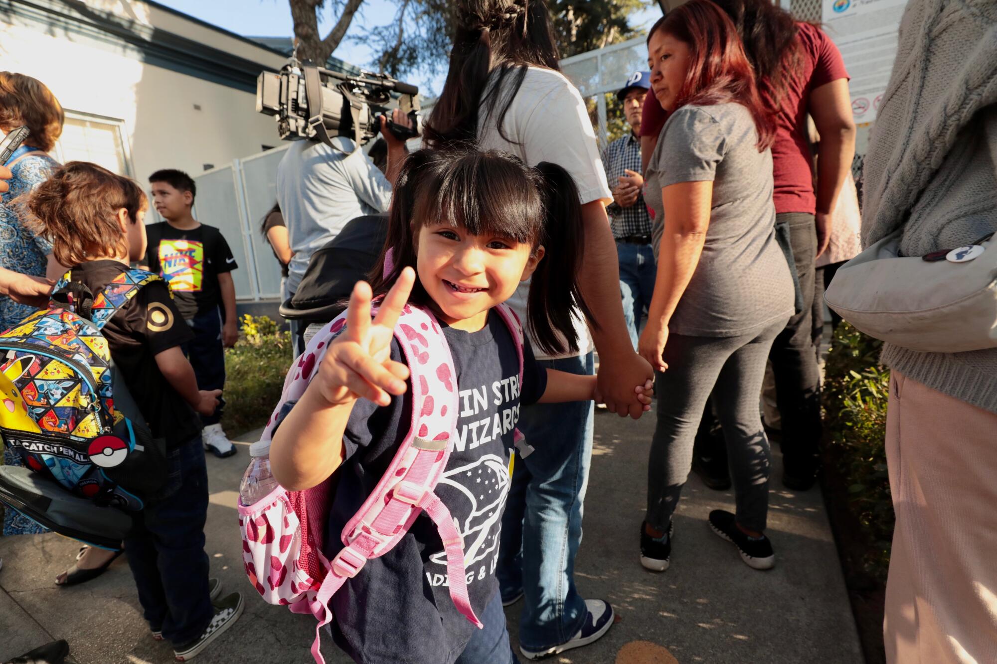 Students with backpacks and their parents at school. One girl holds up her fingers in the peace sign.