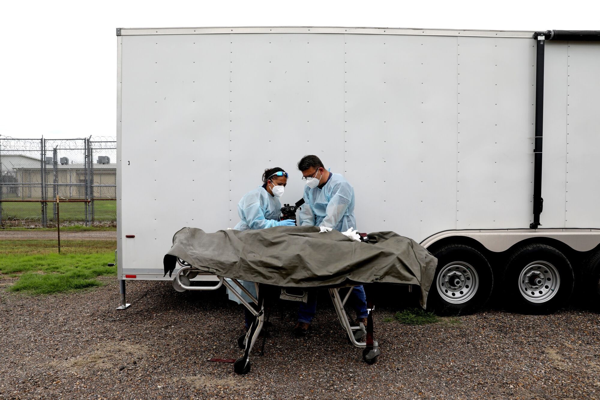 Two forensics workers stand over a body on a gurney beside a trailer