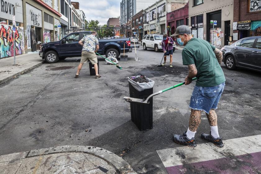 Workers from a Capitol Hill property management company clean up the intersection of 11th and E. Pike Street, Thursday, July 23, 2020, in Seattle, where clothing from the business, Rove, at left was looted and lit on fire in the street last night. Authorities say a group of about 150 people broke windows, stole from businesses and and started a pair of fires in Seattle’s Capitol Hill neighborhood Wednesday night. (Steve Ringman/The Seattle Times via AP)