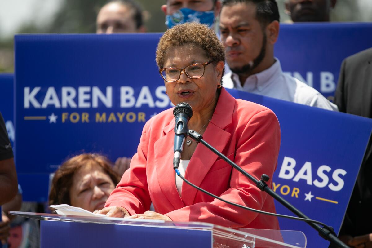 A woman in a pant suit speaks into a microphone. Behind her people hold signs reading "Karen Bass for Mayor."