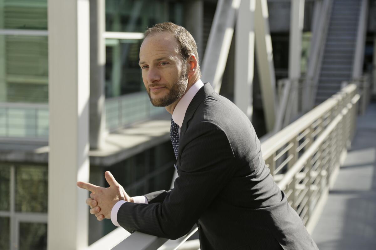 A man poses for a photo leaning his arms on an outdoor railing with a building complex in the background.