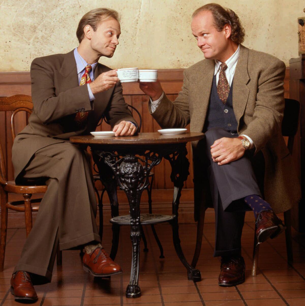 David Hyde Pierce, left, and Kelsey Grammer, both wearing suits and ties, sitting at a table and toasting with tea cups