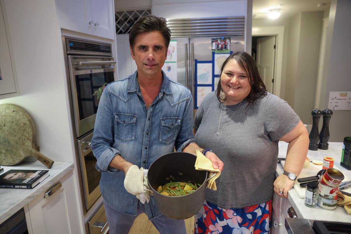 John Stamos stands holding a pot in front of him with Vanessa Franko standing next to him.