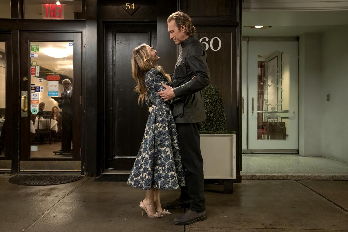 Carrie, in a midi floral dress, hugs Aidan, in a dark jacket, at the waist as she looks up at him.