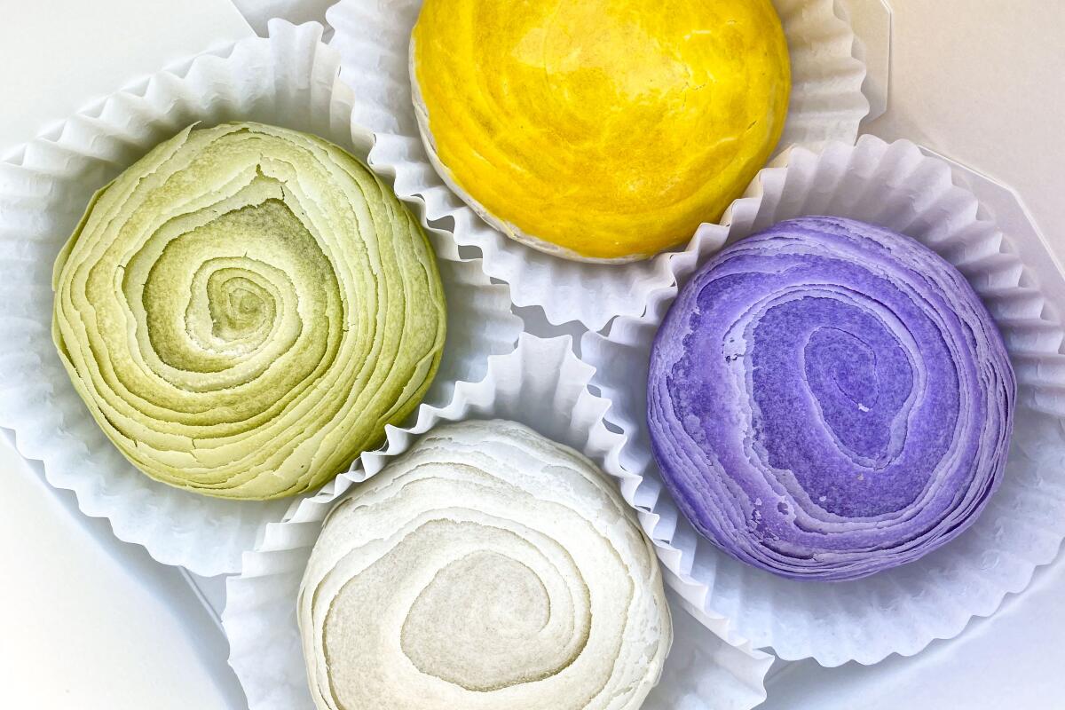 Four round cakes — purple, gold, pale green and white — in white ruffled paper cups