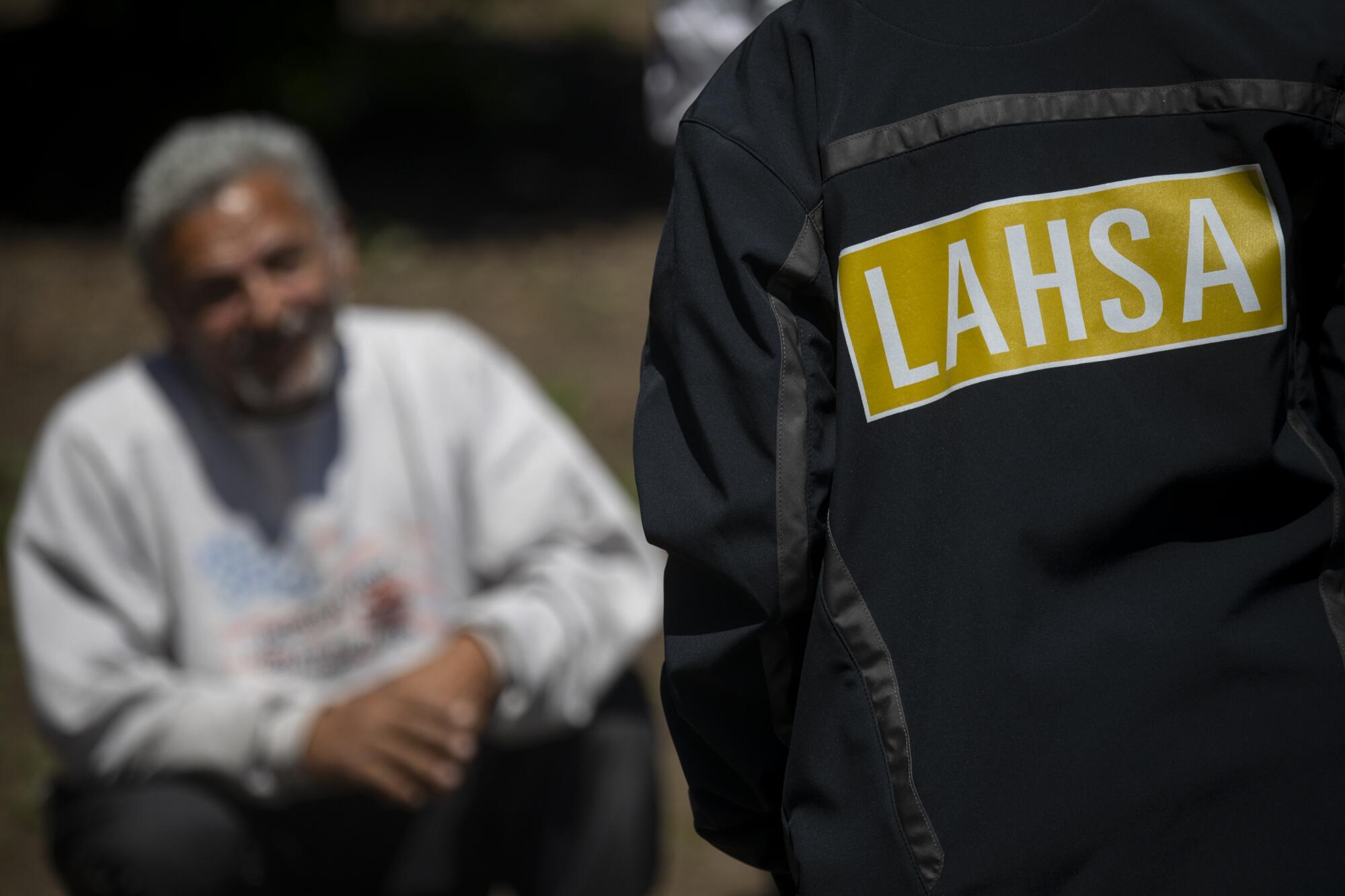 A person in a jacket labeled "LAHSA" stands in the foreground talking to a man in the background.
