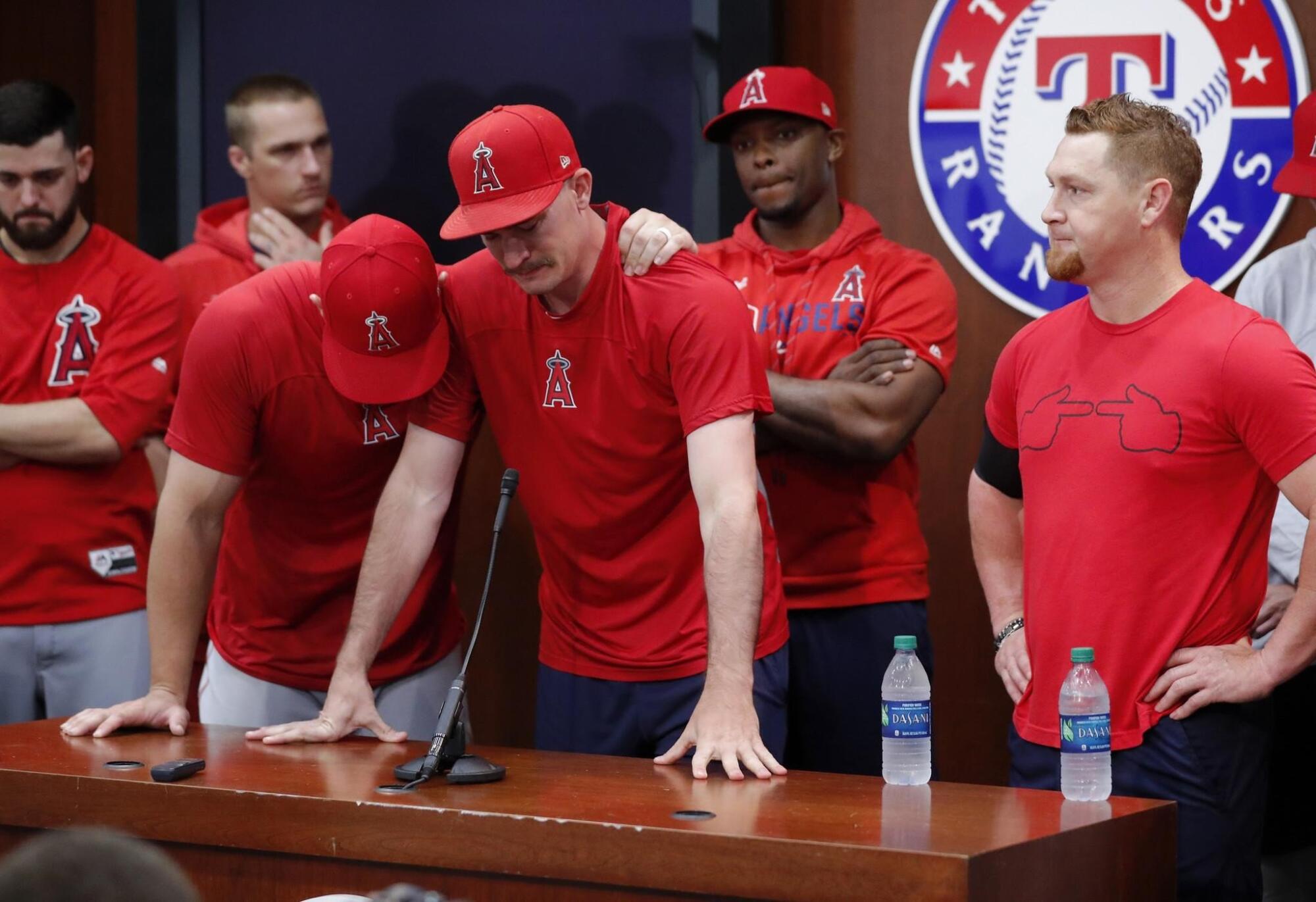 Mike Trout, left, embraces Andrew Heaney at a desk with somber Angels teammates and the Texas Rangers logo behind them
