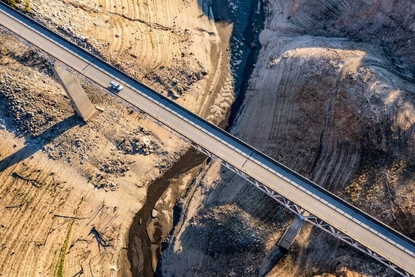 An aerial view shows, at a distance, a vehicle moving along a narrow bridge over brown earth.