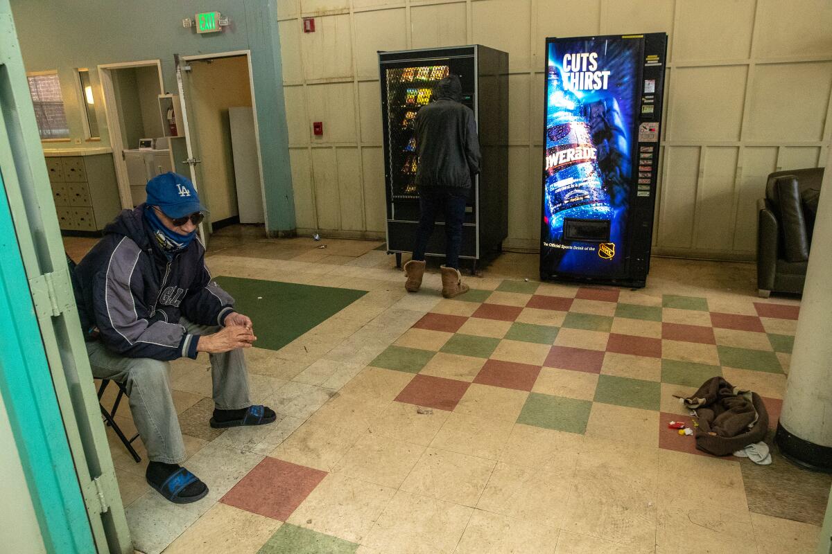 A man sits in a folding chair on a dingy red- and green-checkered floor. Two vending machines stand against a wall.