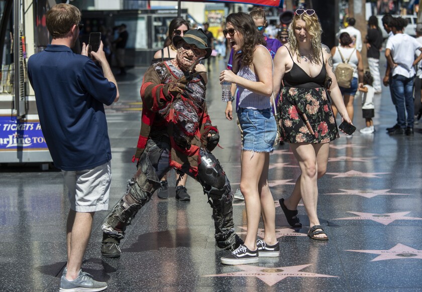 A photo of a man taking a picture of a woman posing with a street performer with people walking behind