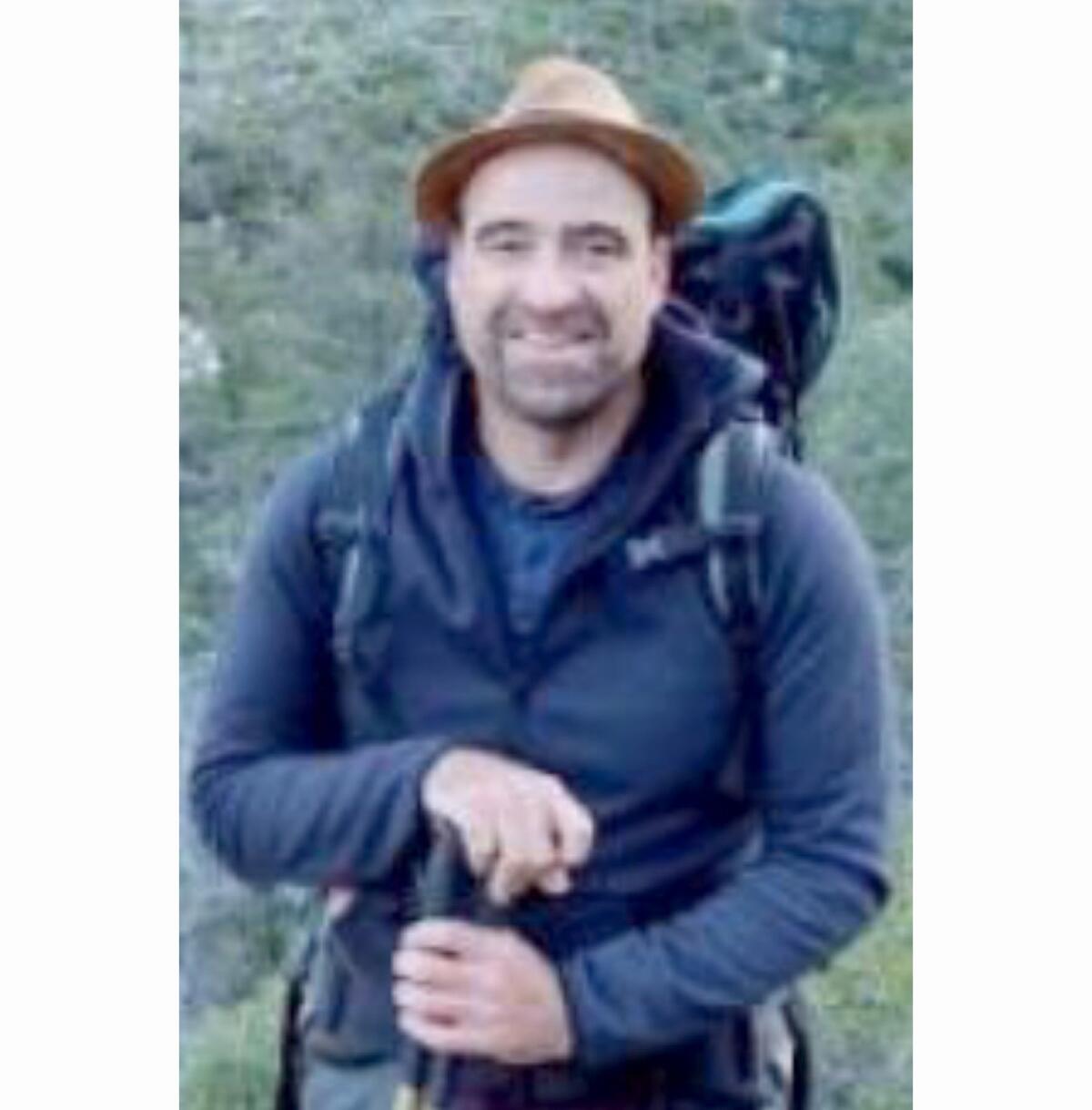 A vertical, low-resolution frame of a smiling man wearing a hat and a backpack. 