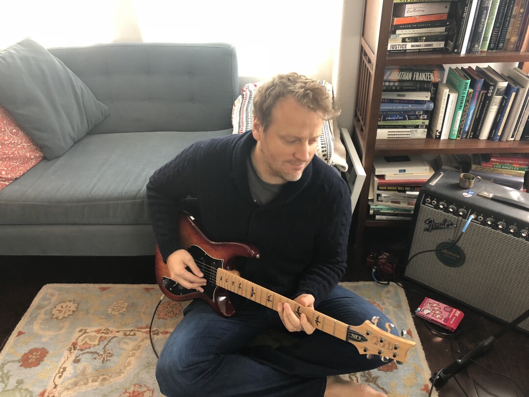 A man plays guitar while seated on a rug.