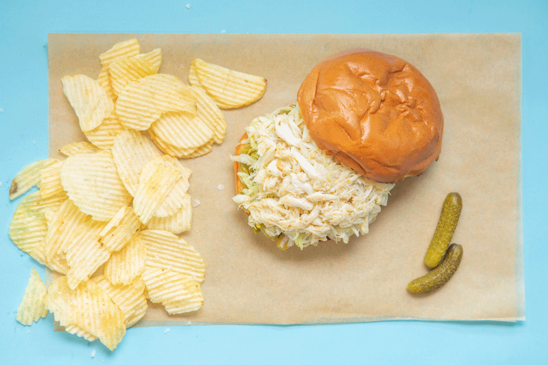 gif of hands picking up a crab sandwich cut in half, with potato chips and gherkins on the side