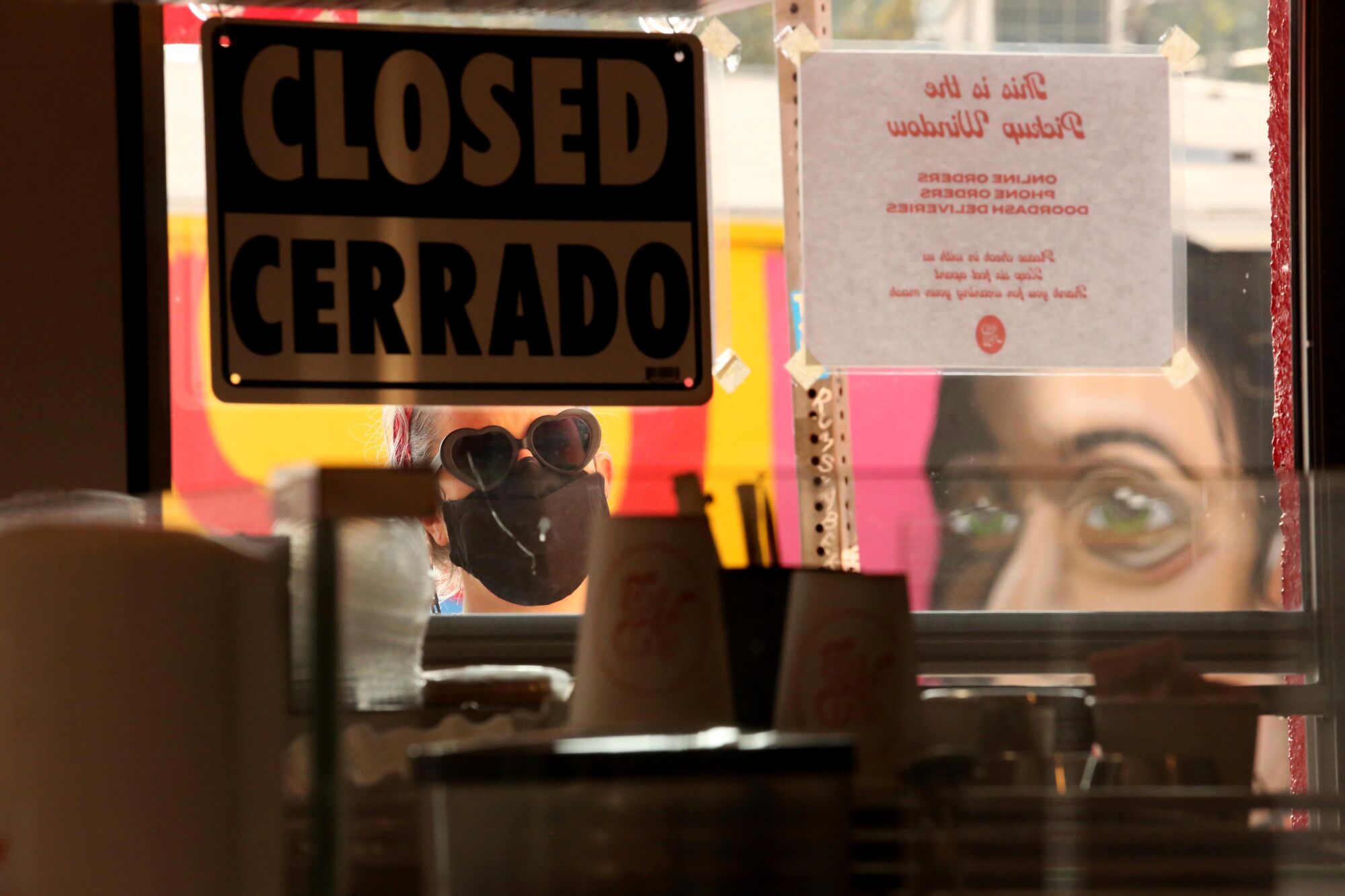A woman in a mask and heart-shaped sunglasses peers into the window of a restaurant, seen from inside.