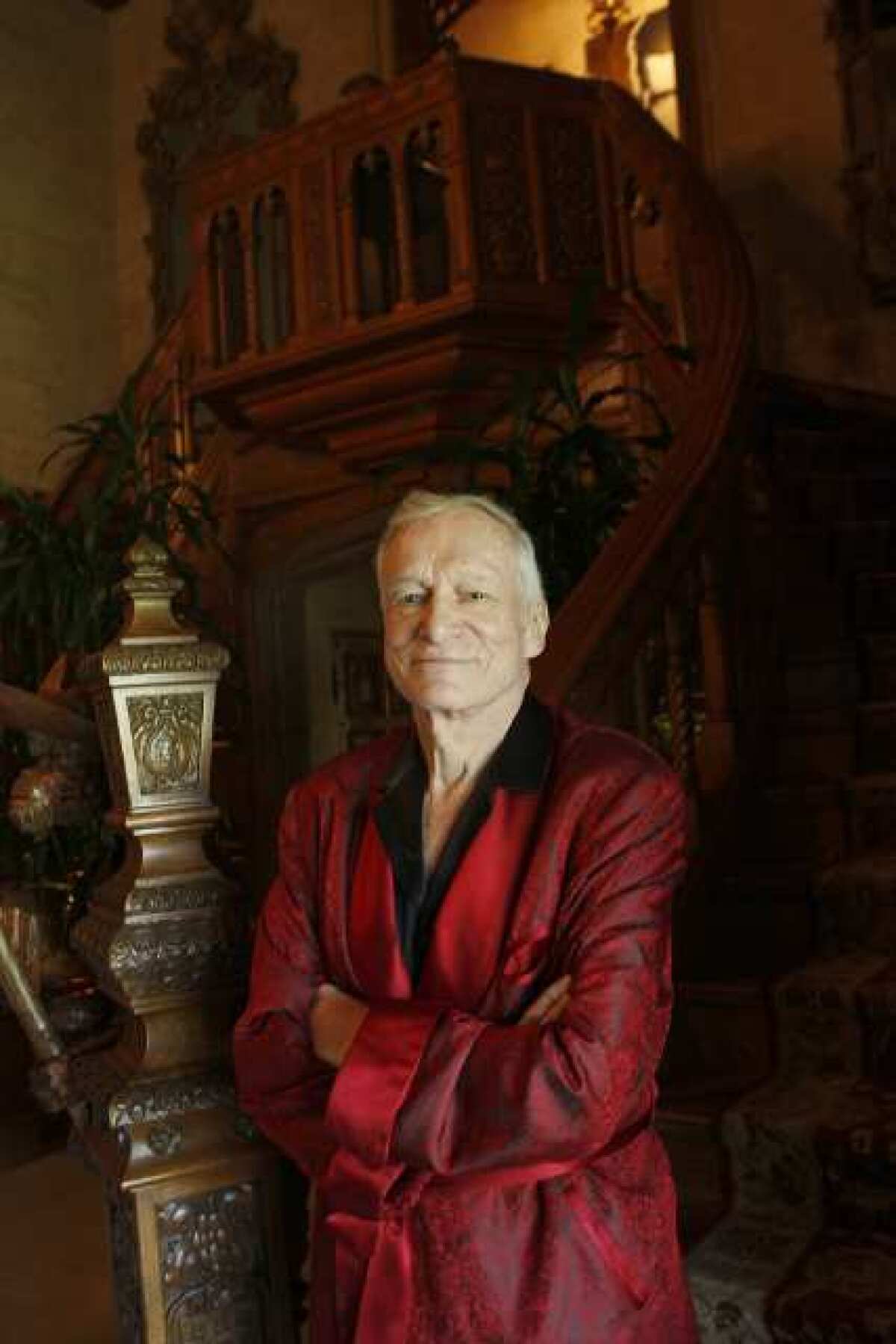 Hugh Hefner standing in a red robe at the foot of a staircase.
