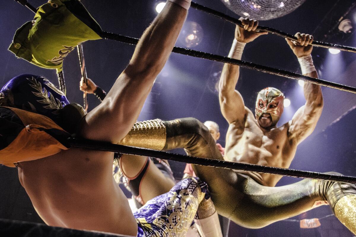A masked Mexican wrestler uses his shiny, shiny boot to pin his opponent against a ring post